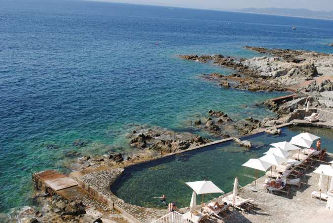A stone sea pool with umbrellas on a rocky Mediterranean coastline.