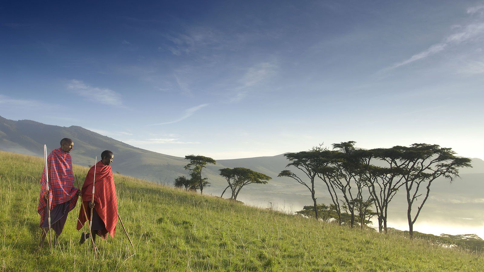 Two Maasai guides walking through green hills at sunrise.